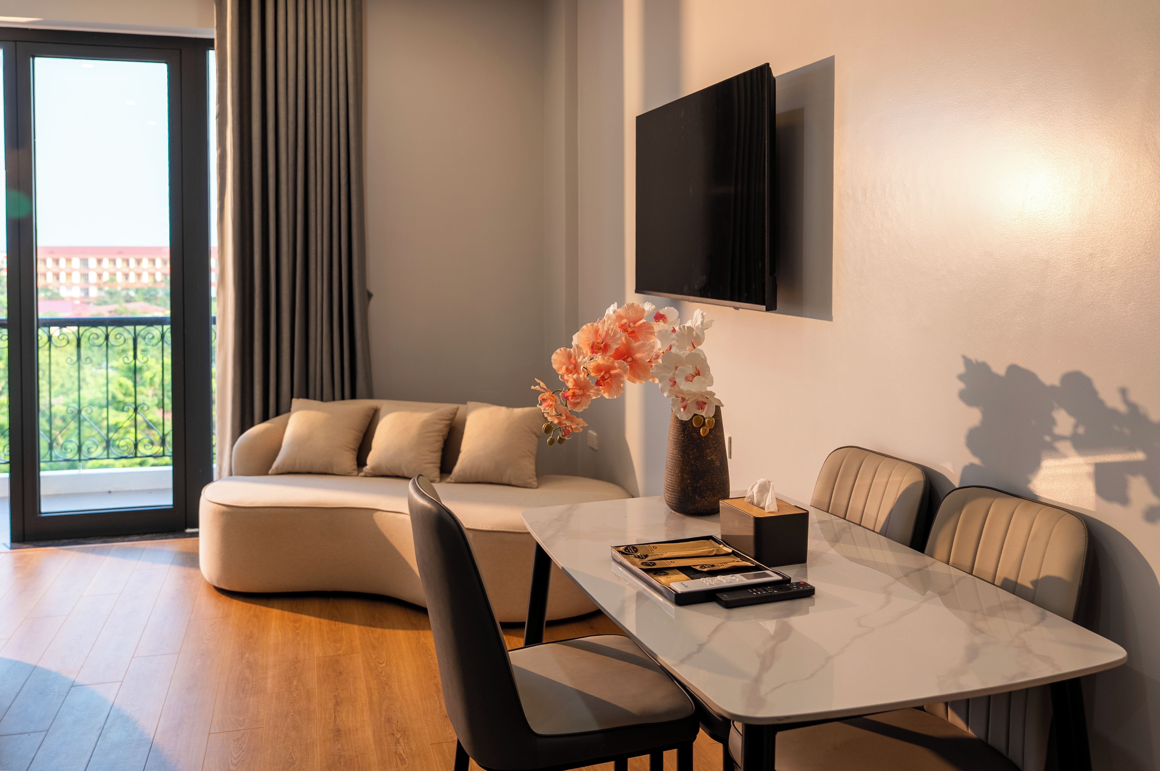 Een moderne woonkamer met een bank, een eettafel met stoelen, een flatscreen televisie aan de muur en een balkon met gordijnen.