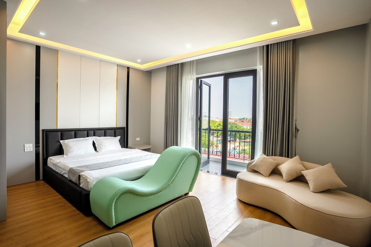 A modern hotel room with a bed, a green armchair, a sofa, and a balcony with a view.