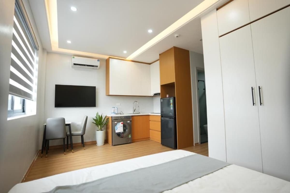 Un dormitorio moderno con cama, escritorio con sillas, lavadora, refrigerador y televisor montado en la pared.