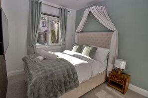 Ein Schlafzimmer mit Bett, Vorhängen, einem Fenster, einem Nachttisch mit Lampe und einem an der Wand befestigten Fernseher.