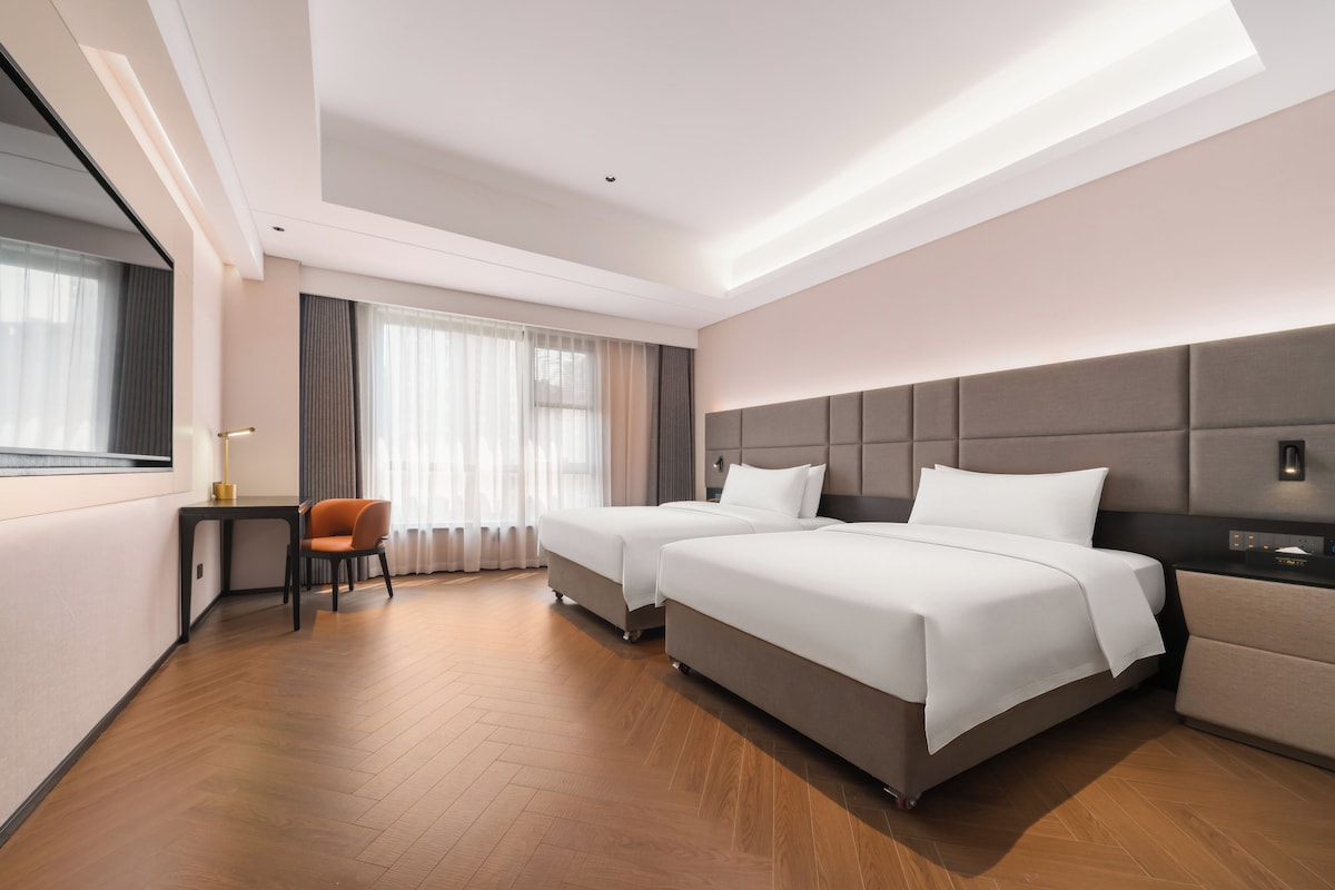 A hotel room with two beds, a desk, a chair, and a large window with curtains.