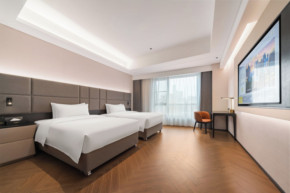 A modern hotel room with two beds, a desk, a chair, and a large window with curtains.