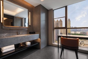 A modern bathroom with a large mirror, a vanity with a sink, and a chair by a window with a city view. - Howard Johnson Club Hotel Wuhan Jiyuqiao (Wuhan)