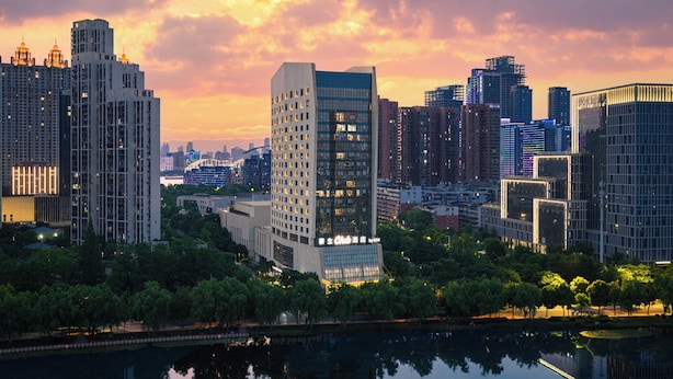 Paisagem urbana com edifícios modernos e altos, um corpo d'água em primeiro plano e um céu ao pôr do sol.