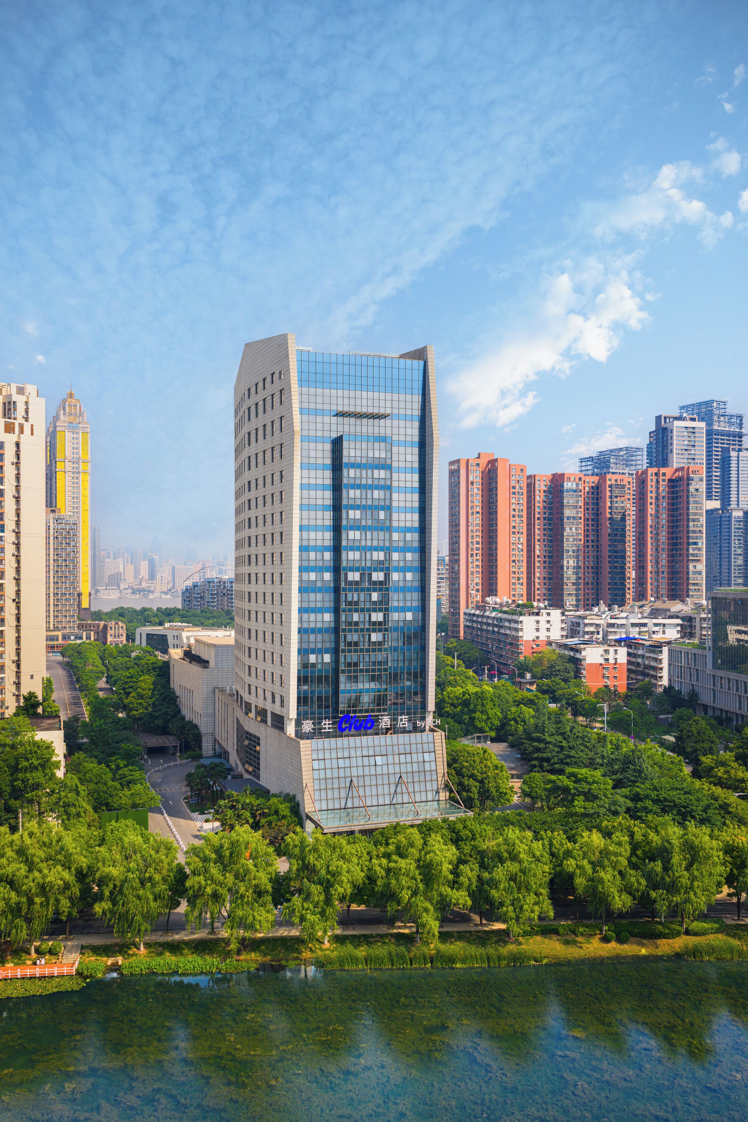 A modern hotel with a glass facade, surrounded by greenery and a body of water.
