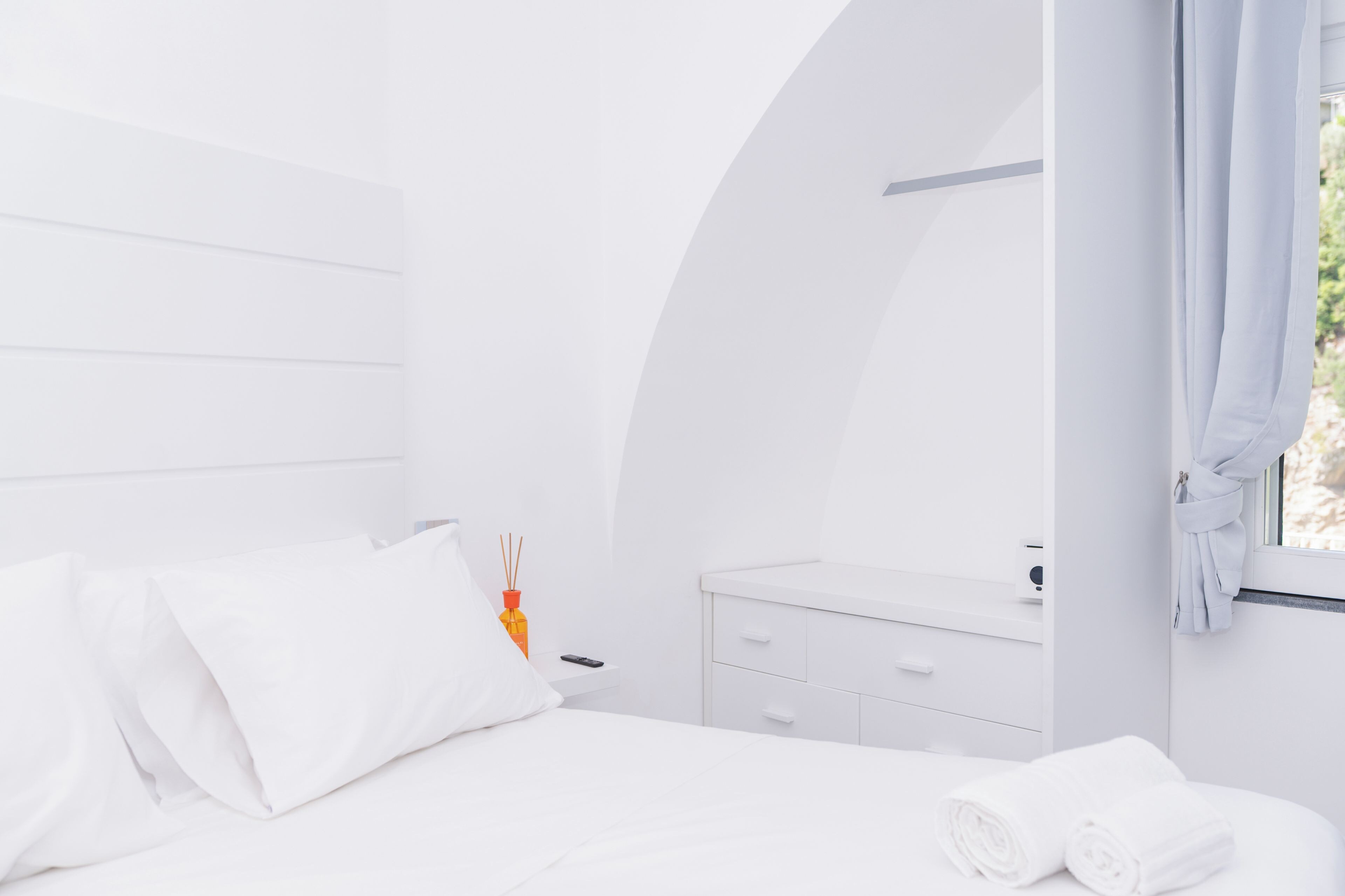 A modern, white-themed bedroom with a bed, a nightstand, a mirror, and a window with curtains.
