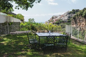 Une terrasse extérieure avec une table et des chaises, entourée d’une clôture blanche, donnant sur une colline parsemée de bâtiments.