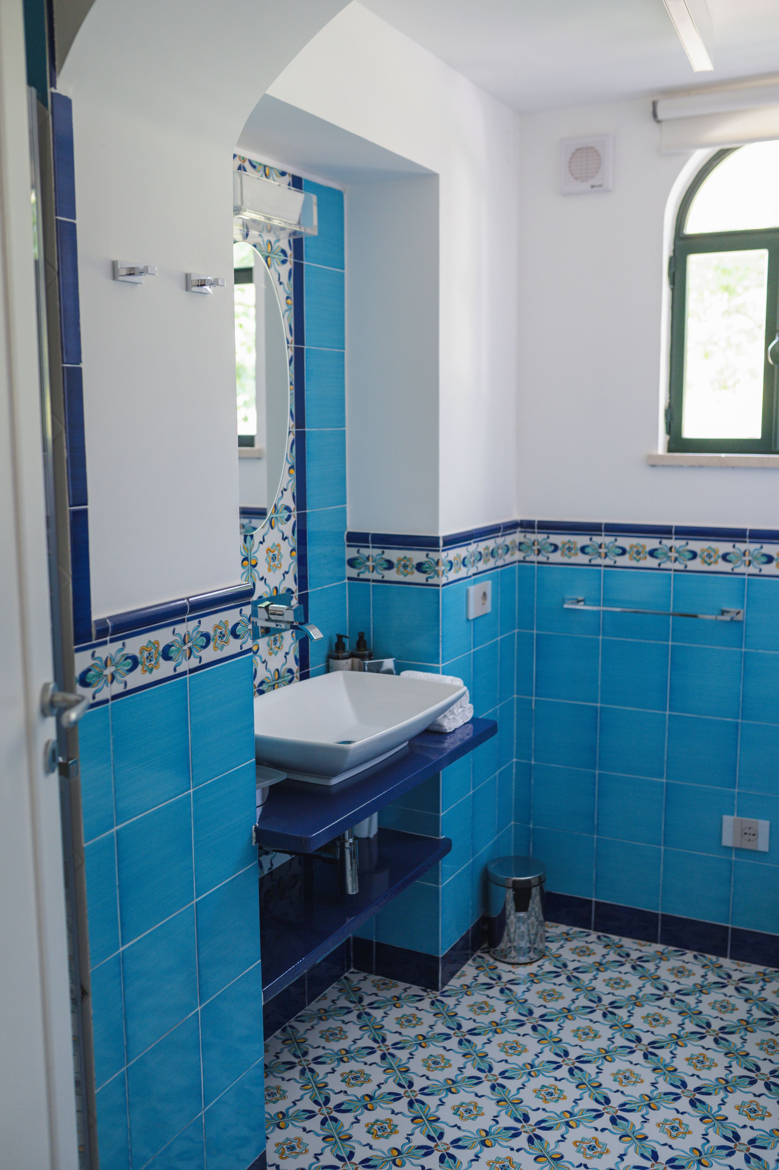 Une salle de bain avec un lavabo blanc, des carreaux bleus et une fenêtre.