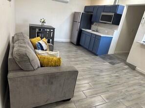 A compact living space with a sofa, a small table with a vase, and a kitchenette area with a refrigerator, microwave, and cabinets. - Cozy  studio in welcoming Indio with AC (Indio)