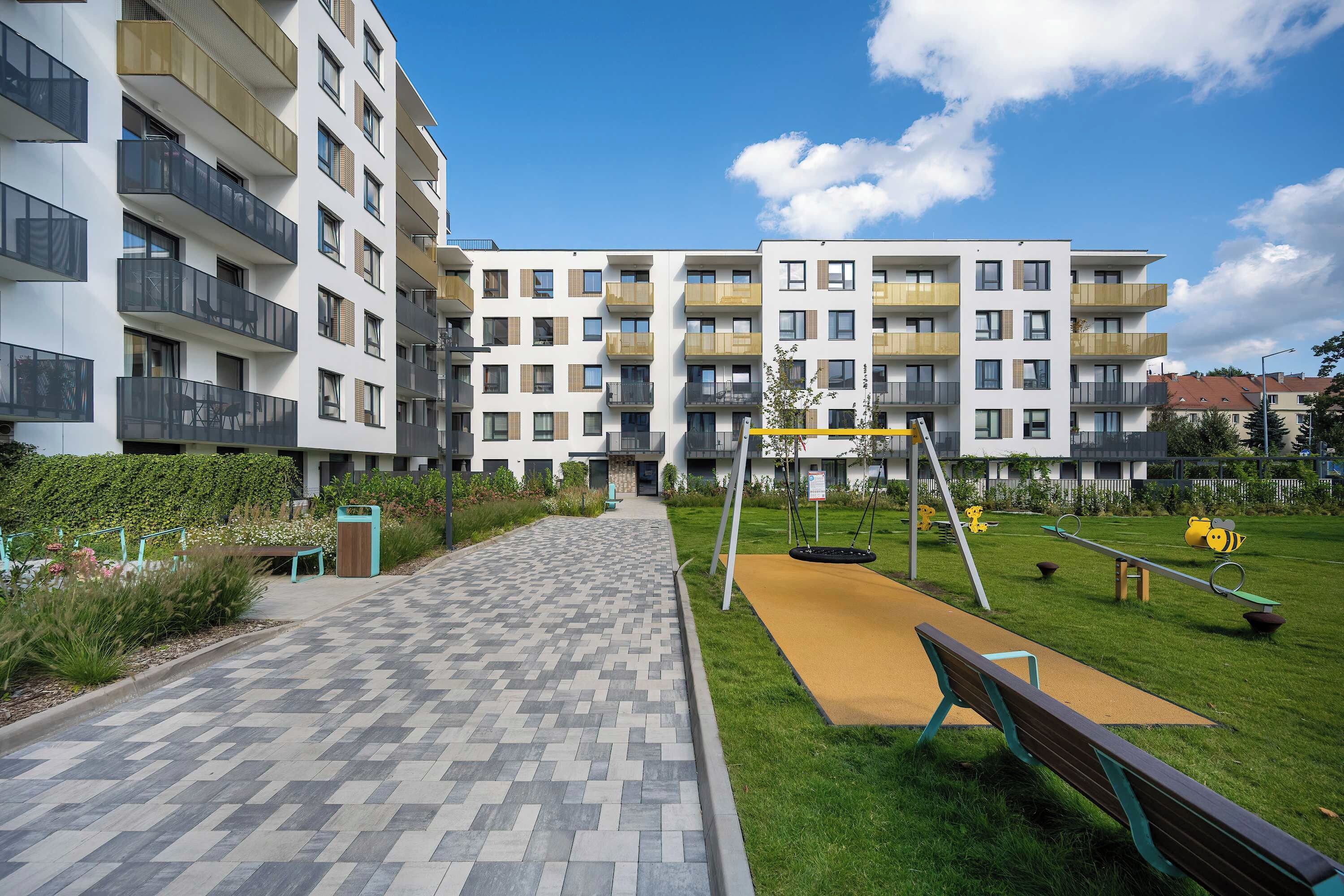 Ein Wohngebäude mit mehreren Balkonen, einem gepflasterten Weg und einem Spielplatz mit Schaukeln und einer Wippe.