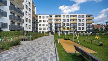 Ein Wohngebäude mit mehreren Balkonen, einem gepflasterten Weg und einem Spielplatz mit Schaukeln und einer Wippe.