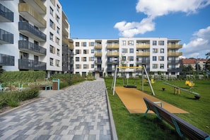 Ein Wohngebäude mit mehreren Balkonen, einem gepflasterten Weg und einem Spielplatz mit Schaukeln und einer Wippe.