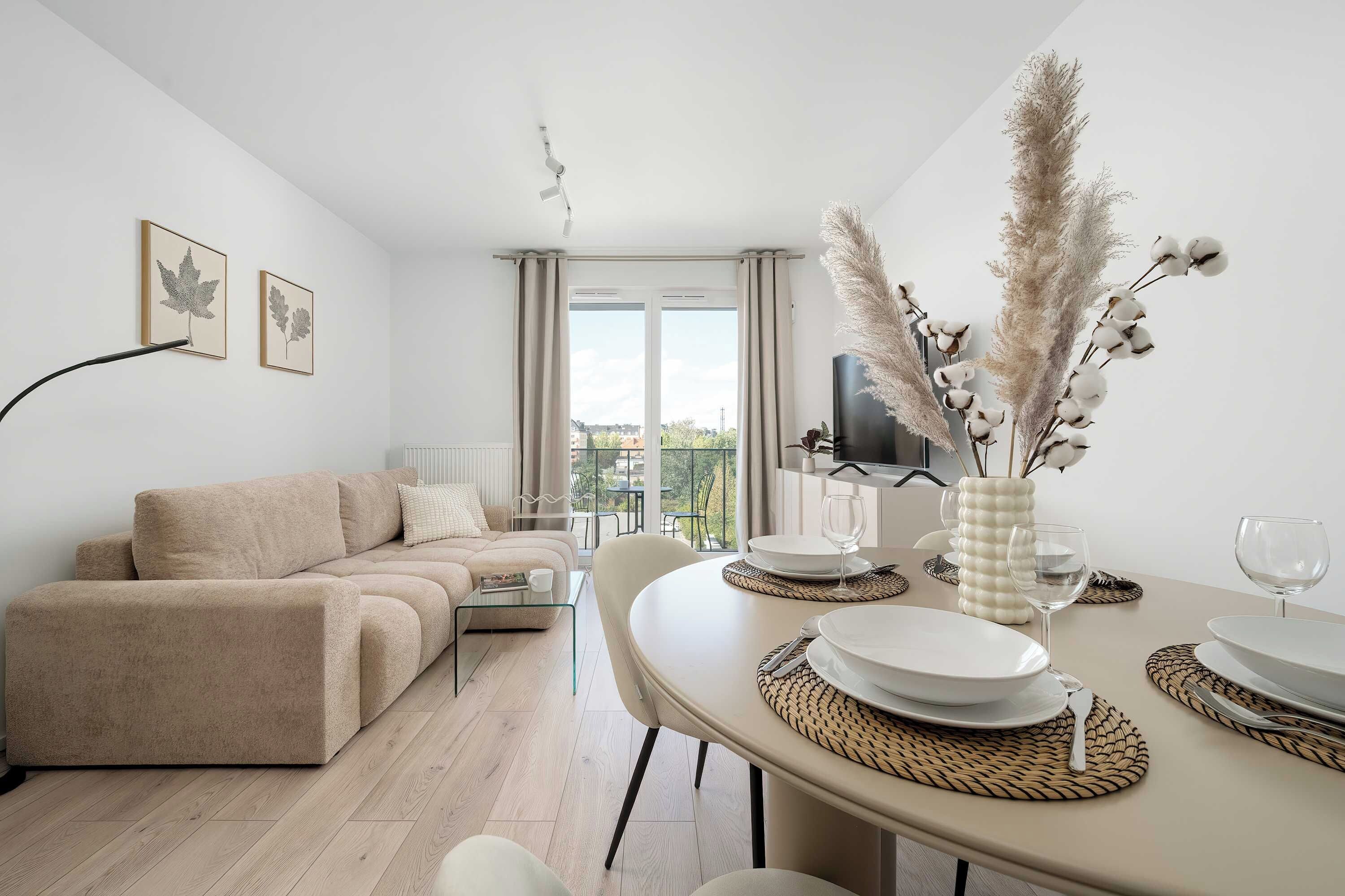 Ein modernes Wohnzimmer mit einem beigefarbenen Sofa, einem Esstisch mit Tellern und Gläsern sowie einem großen Fenster mit Vorhängen.