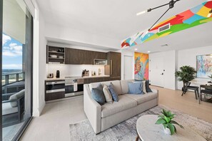 Cozinha moderna com eletrodomésticos embutidos, área de jantar com mesa redonda e sala de estar com sofá e planta.
