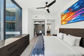 Quarto de hotel moderno com uma cama grande, ventilador de teto, vista por uma porta de vidro e um quadro colorido na parede.