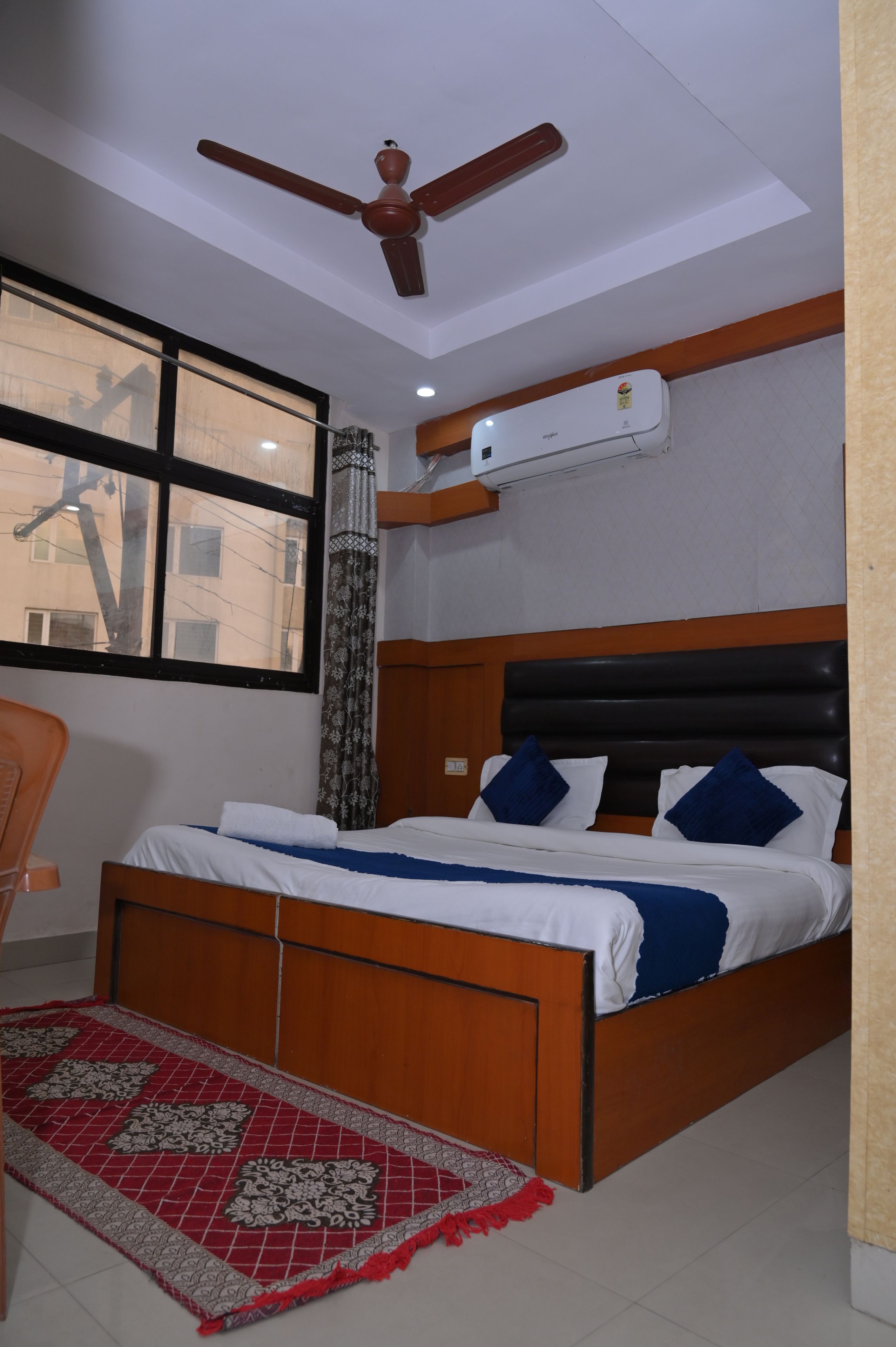 Un dormitorio con cama, ventilador de techo, equipo de aire acondicionado, ventana y alfombra estampada.