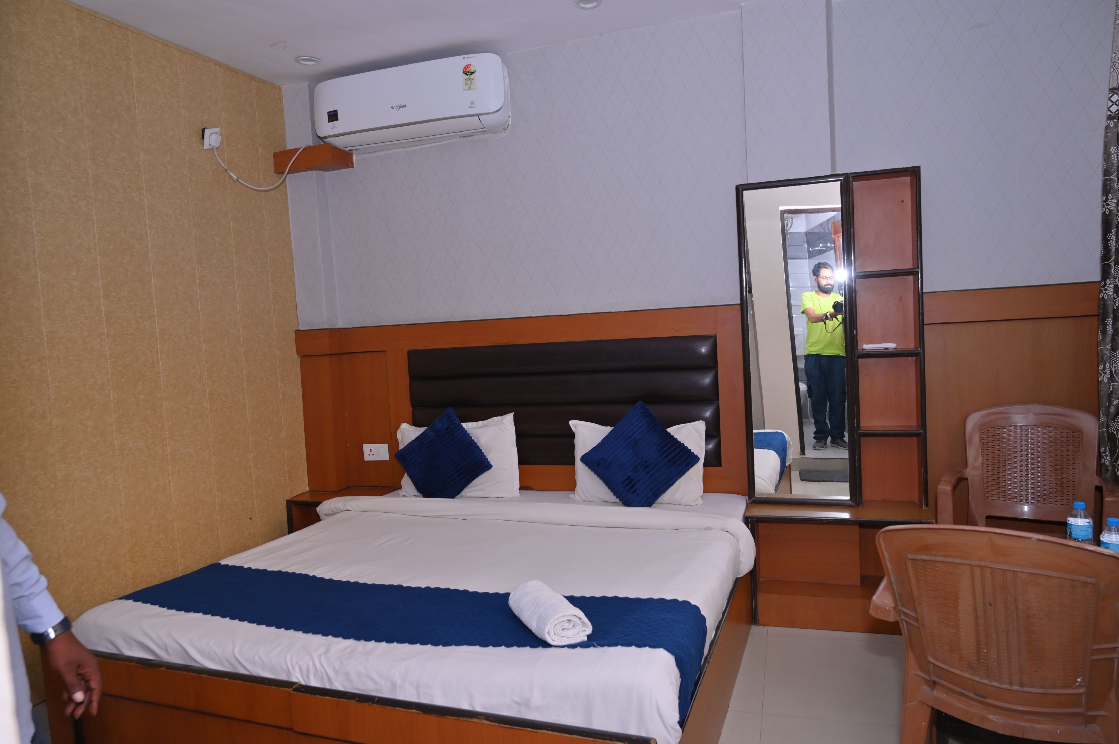 Una habitación de hotel con cama, mesita de noche, silla, aire acondicionado y espejo.
