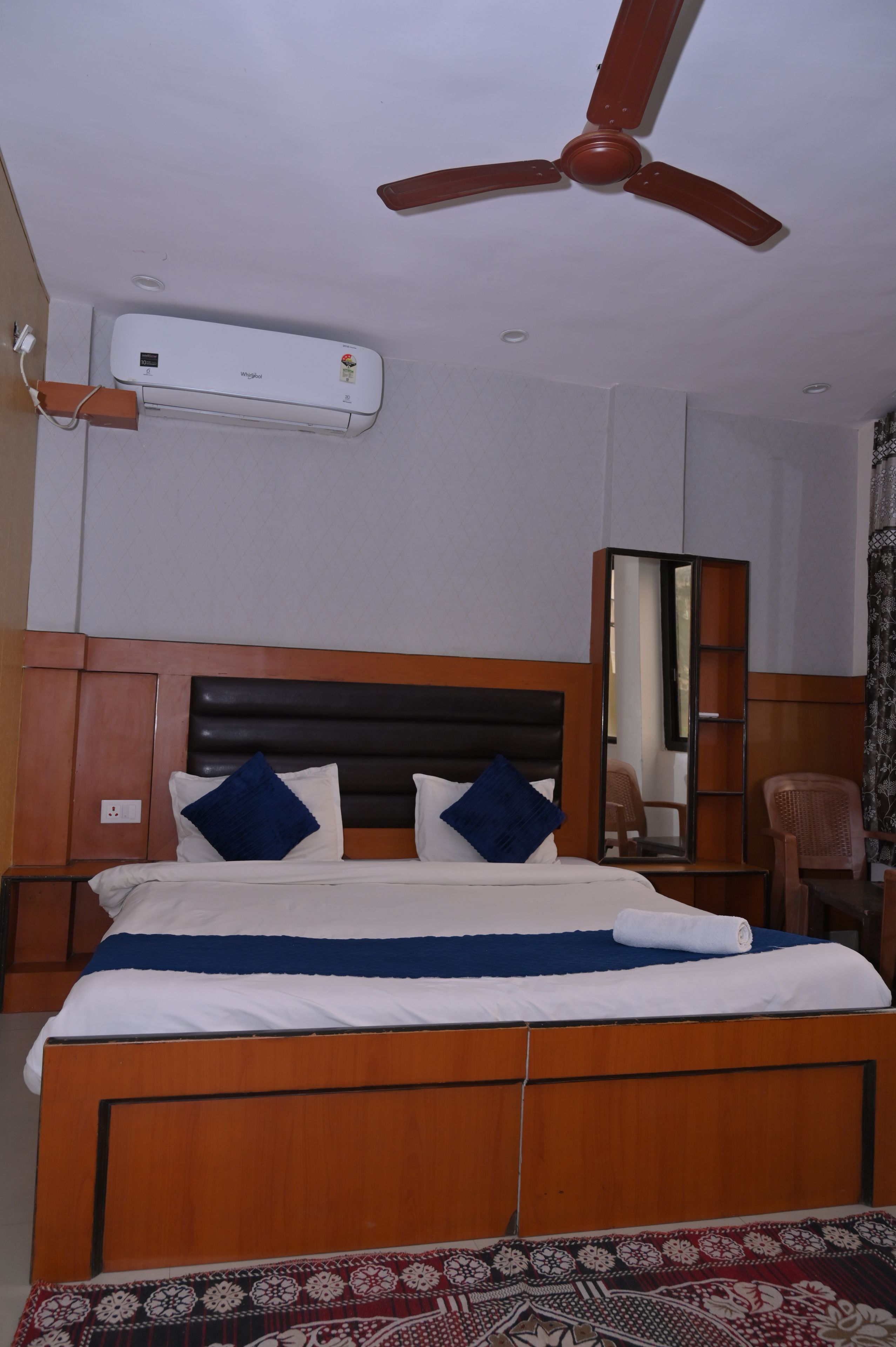 Un dormitorio con cama, ventilador de techo, equipo de aire acondicionado, espejo, silla y alfombra estampada.