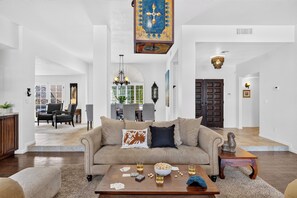 A spacious living room with a beige sofa, a coffee table with cards and drinks, and a dining area in the background. - Private Pool + Hot Tub + Fire Pit + Covered Patio | Mins to Golf | Casa Talavera (Scottsdale)