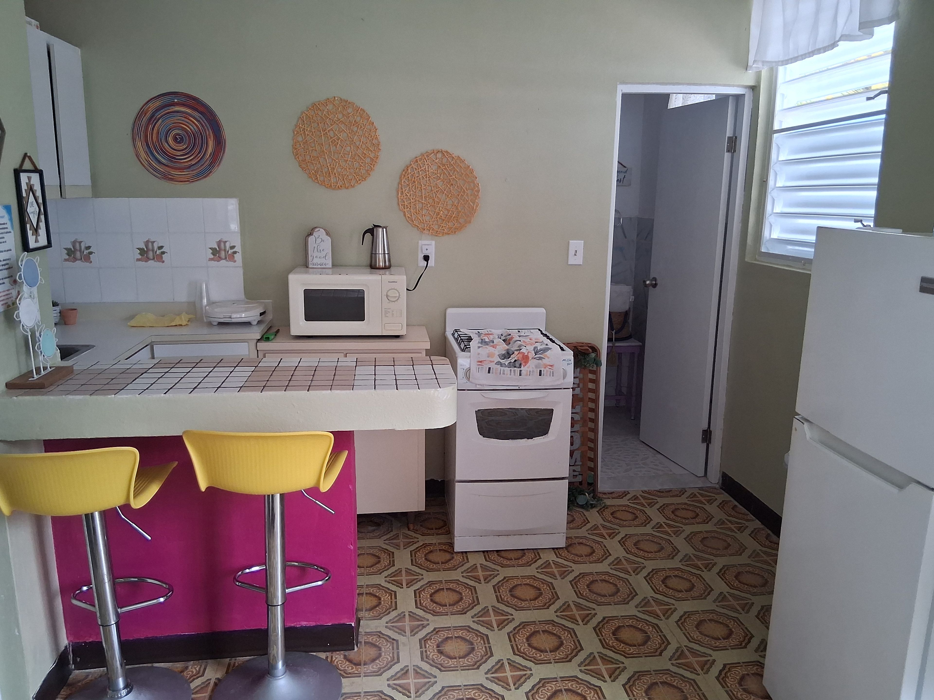 Cocina compacta con microondas, un refrigerador pequeño y un lavamanos. Hay dos sillas amarillas junto a una barra pequeña.