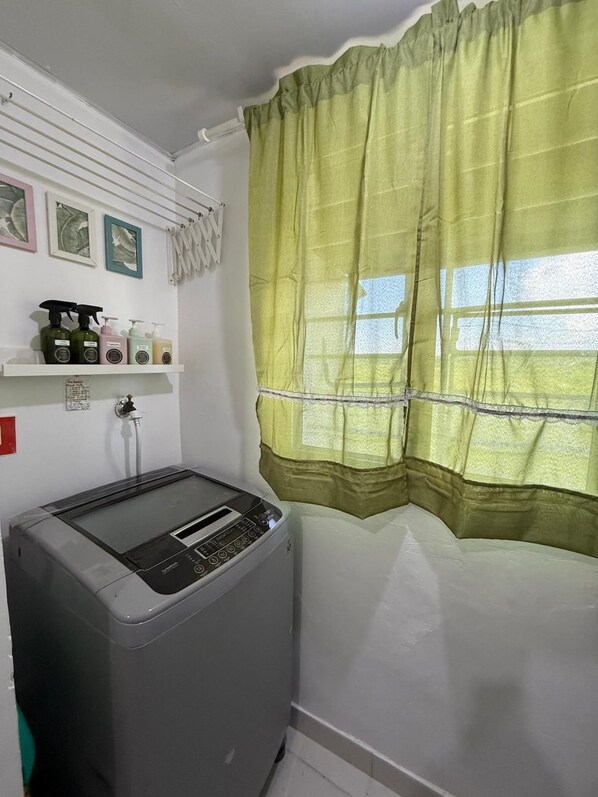 Ein Waschraum mit einer großen Waschmaschine, einem Regal voller Reinigungsmittel und einem Fenster mit grünem Vorhang.