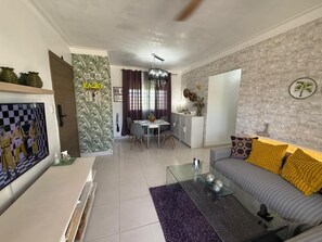 Ein Wohnzimmer mit einer Couch, einem Couchtisch und einem Fernseher. Es gibt eine Essecke mit Tisch und Stühlen. Der Raum hat eine Ziegelwand und einen Deckenventilator.