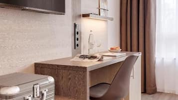 A modern hotel room with a wooden desk, a chair, a flat-screen TV mounted on the wall, and a suitcase on the floor.