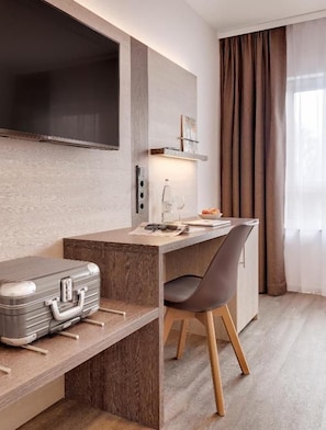Une chambre d’hôtel moderne avec un bureau en bois, une chaise, un téléviseur à écran plat fixé au mur et une valise posée sur le sol.