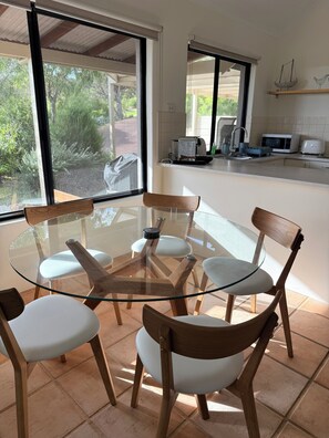 A modern kitchen with a glass dining table surrounded by wooden chairs, large windows, and a view of greenery outside.
