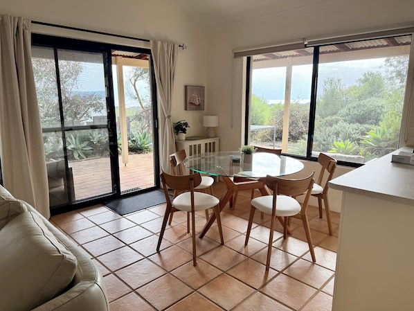 A dining area with a glass table and wooden chairs, a white sofa, and large windows with a view of greenery.