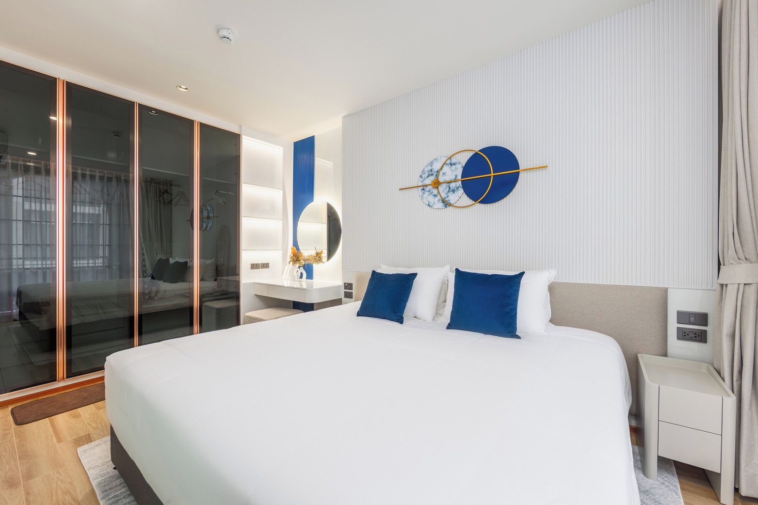 Une chambre d’hôtel moderne avec un grand lit, une cloison en verre, une table de chevet et une horloge fixée au mur.