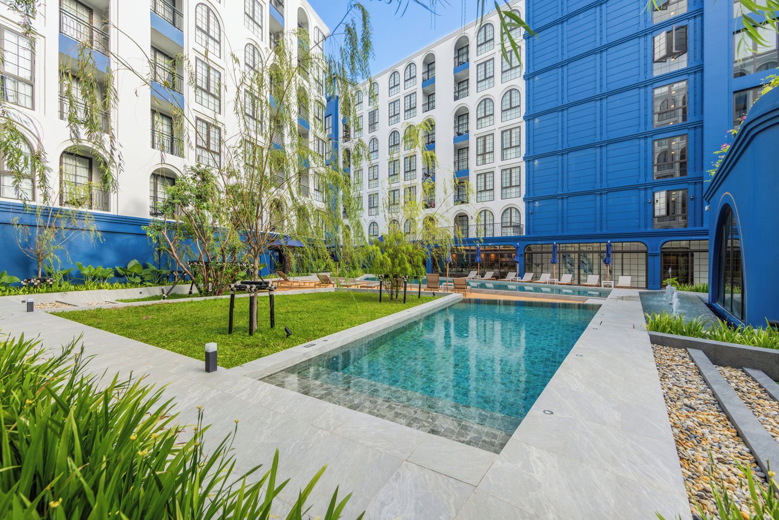 Un patio de hotel con piscina, sillas de descanso y un edificio azul con ventanas arqueadas.