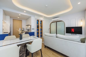 A modern living room with a sofa, a dining table with chairs, a TV mounted on the wall, and a bookshelf.
