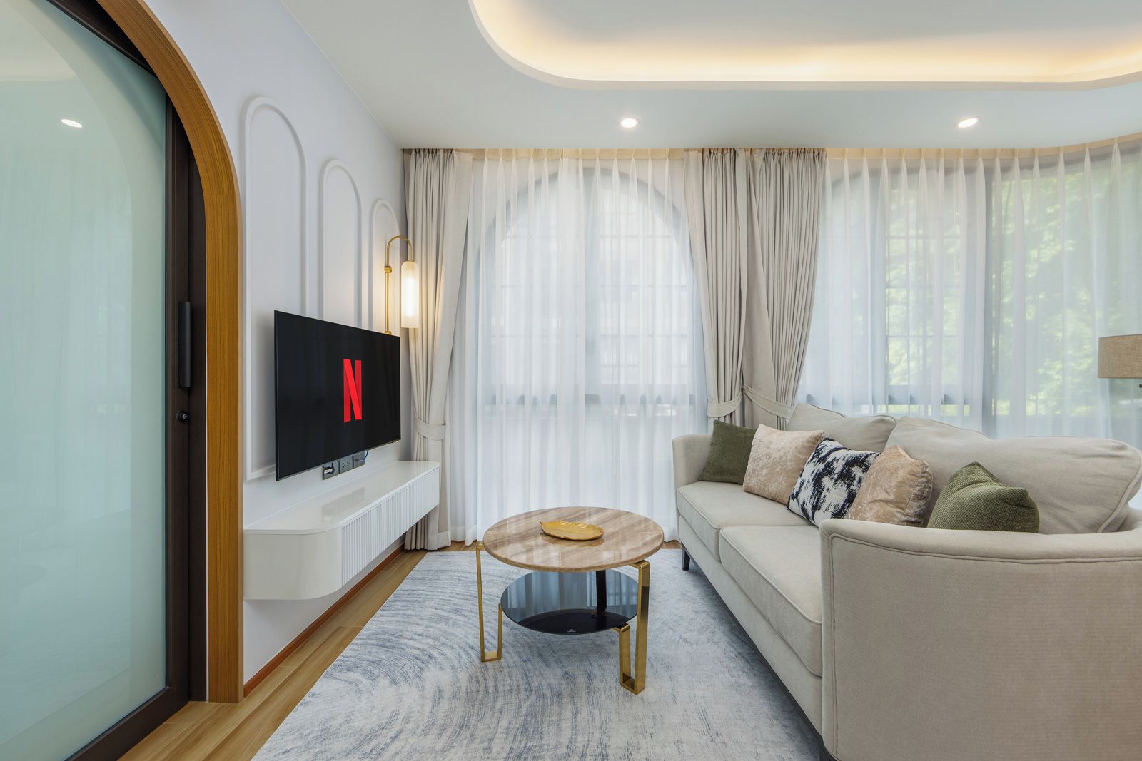 Una sala moderna con un televisor de pantalla plana mostrando el logo de Netflix, un sofá color beige con cojines estampados, una mesa de centro redonda y cortinas translúcidas.