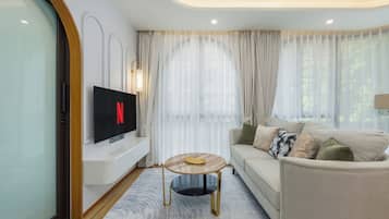 Una sala moderna con un televisor de pantalla plana mostrando el logo de Netflix, un sofá color beige con cojines estampados, una mesa de centro redonda y cortinas translúcidas.
