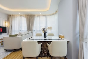 A modern living room with a glass-top dining table, two white chairs, a sofa, a TV, and a vase with flowers.