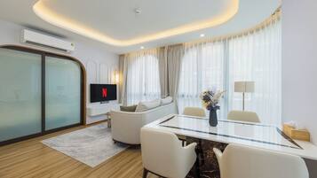 Una habitación de hotel moderna con una mesa de comedor de vidrio, un sofá, un televisor y un ventanal con cortinas.