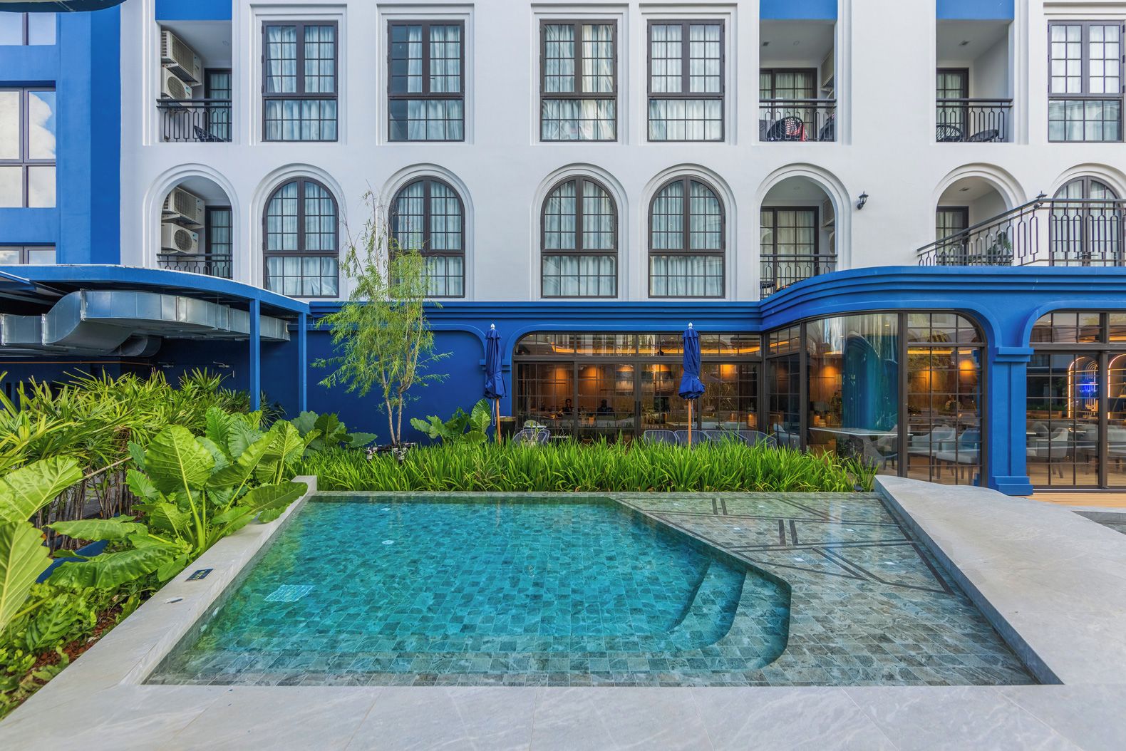 Un patio de hotel con piscina, abundante vegetación y fachada de edificio azul y blanco.