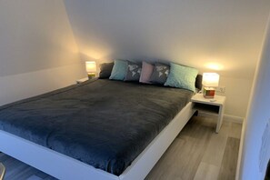 Ein ordentlich bezogenes Bett mit dunkler Bettdecke, zwei Nachttische mit Lampen und ein Kopfkissen mit Weltkarte.