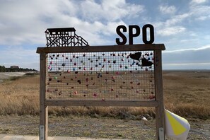 Ein hölzernes Schild mit einer Anordnung von Vorhängeschlössern, einer Struktur mit der Aufschrift 'SPO' oben und einer flachen Landschaft im Hintergrund.