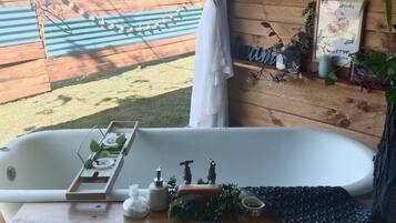 A bathroom with a bathtub, a wooden shelf with plants, and a view of a wooden deck and hills.