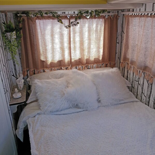 Cama com roupa de cama branca, mesa de cabeceira com abajur e parede com padrão de árvore.