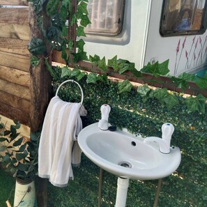 Uma pia branca ao ar livre, com uma torneira, uma toalha pendurada em um gancho e folhagem verde ao fundo.