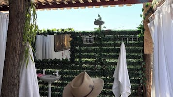 Un espace extérieur abrité, doté d’un banc en bois, d’un arbre et d’un mur où sont accrochés divers objets.