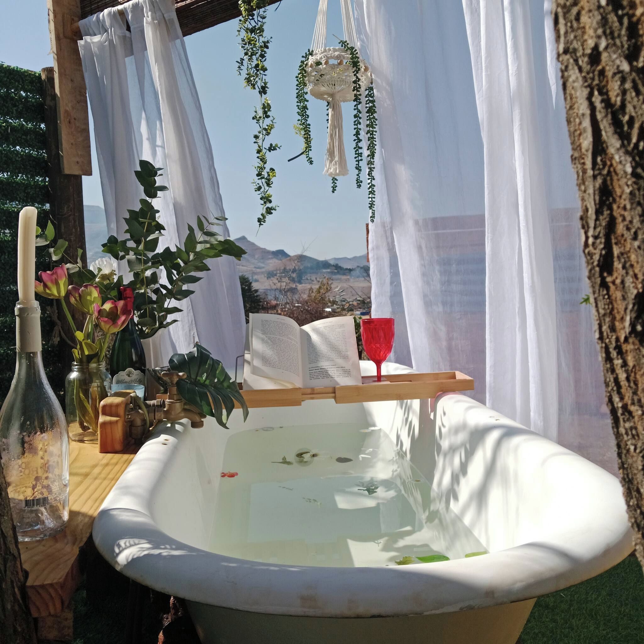 Un décor extérieur rustique avec une baignoire blanche, une table en bois surmontée d’un verre rouge et une décoration en macramé suspendue.