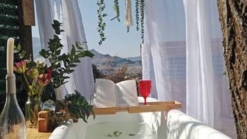 Un décor extérieur rustique avec une baignoire blanche, une table en bois surmontée d’un verre rouge et une décoration en macramé suspendue.
