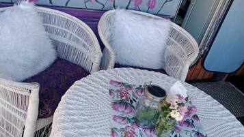 Un rincón acogedor con dos sillones de mimbre blanco, una mesa redonda con mantel floreado y un frasco de vidrio decorativo.