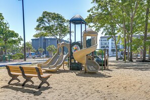 Ein Spielplatz mit Rutschen und einer Bank in einem sandigen Bereich, im Hintergrund Bäume und Gebäude.