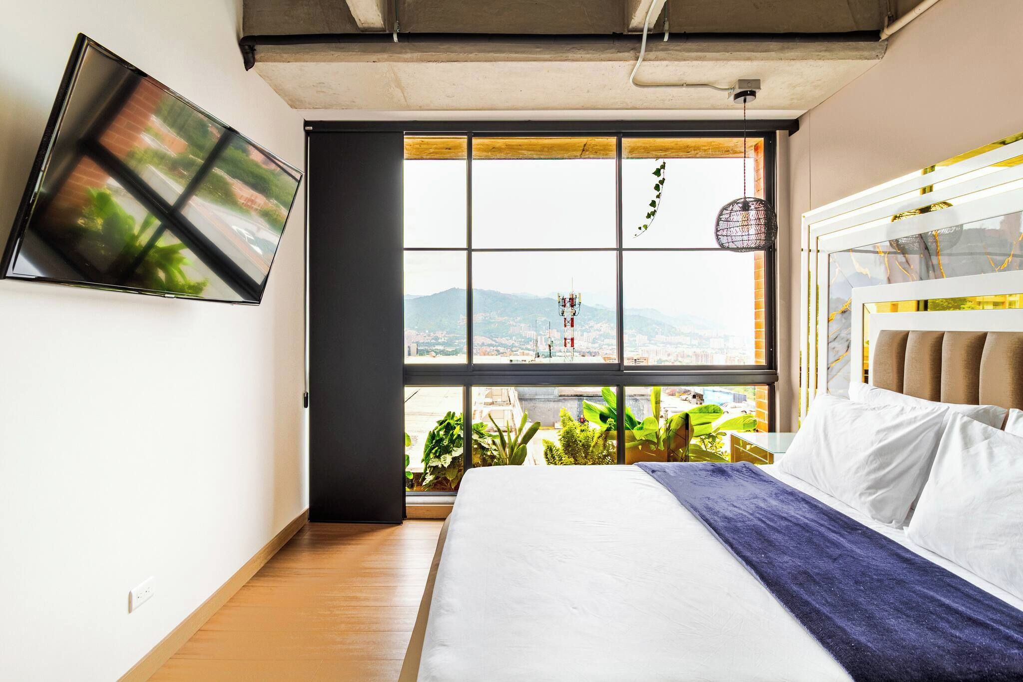 Un dormitorio con cama, televisor montado en la pared y un amplio ventanal con vista a la ciudad.