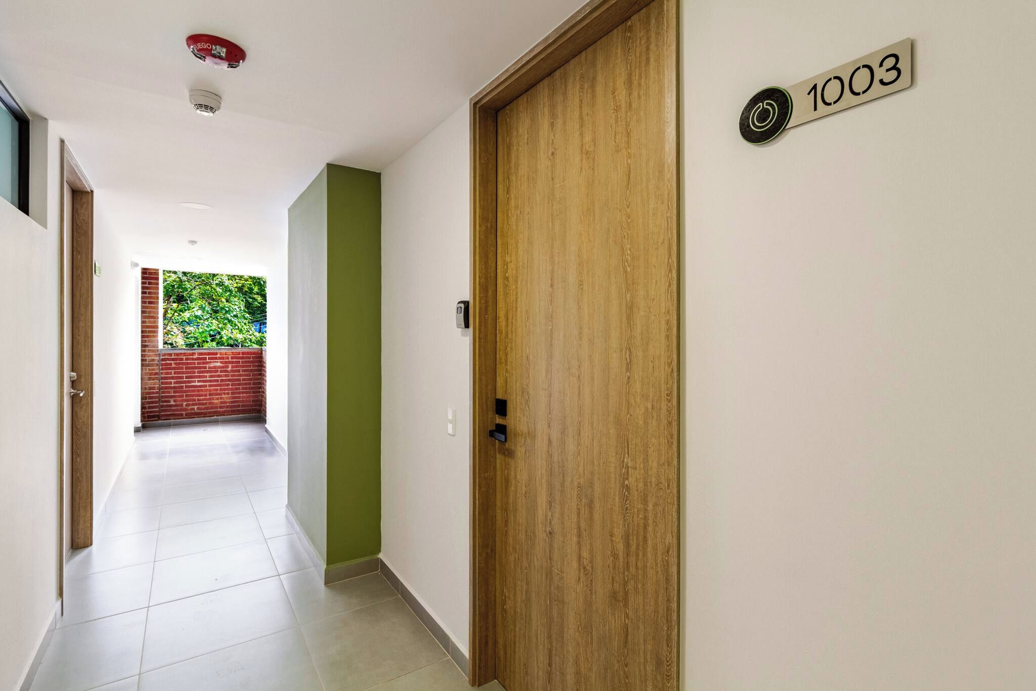 Ein Flur mit einer Holztür mit der Aufschrift '1003', einer grünen Wand und Blick auf eine Ziegelwand und Grünflächen im Freien.